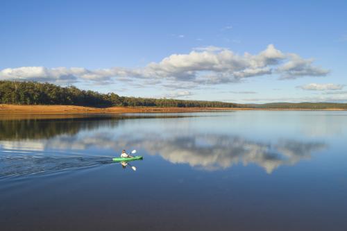 reflections of a blue sky with a few clouds on the still waters of the dam with forest in the background