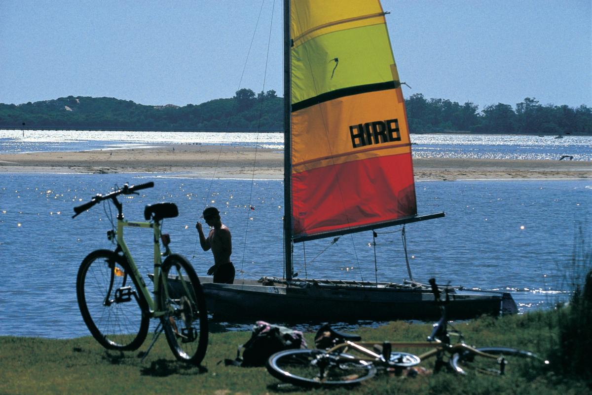 Leschenault Peninsula Conservation Park man stabnding in water near catamaran with orange and red sail and two bicycles nearby on grass